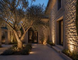 Giardino residenziale al crepuscolo con faretti incassati nel pavimento che illuminano una facciata e un ulivo.
